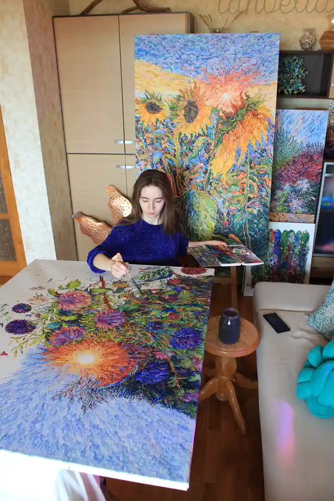 A woman in a blue sweater sits at a table painting a vibrant, textured floral scene with a sunburst. Behind her, a large…