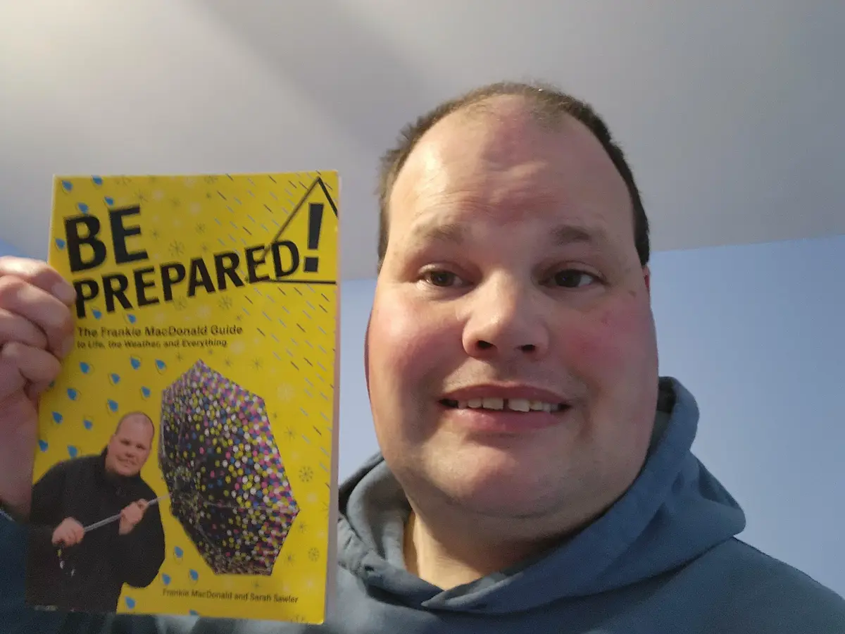 A man in a blue hoodie holds up a yellow book titled 'BE PREPARED! The Frankie MacDonald Guide to Life, the Weather, and…