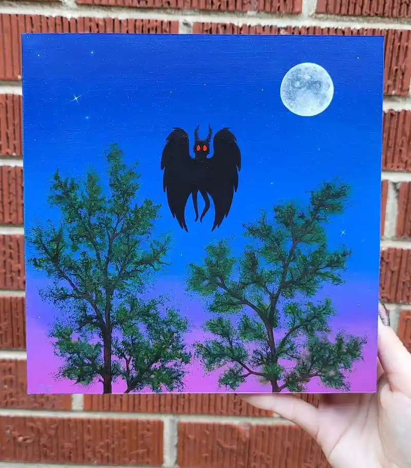 A hand holds a small painting depicting a dark, winged creature with glowing red eyes flying between two green trees against…