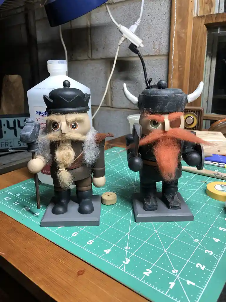 Two handcrafted wooden nutcracker figurines stand on a green cutting mat, one with a crown and grey beard, the other with…