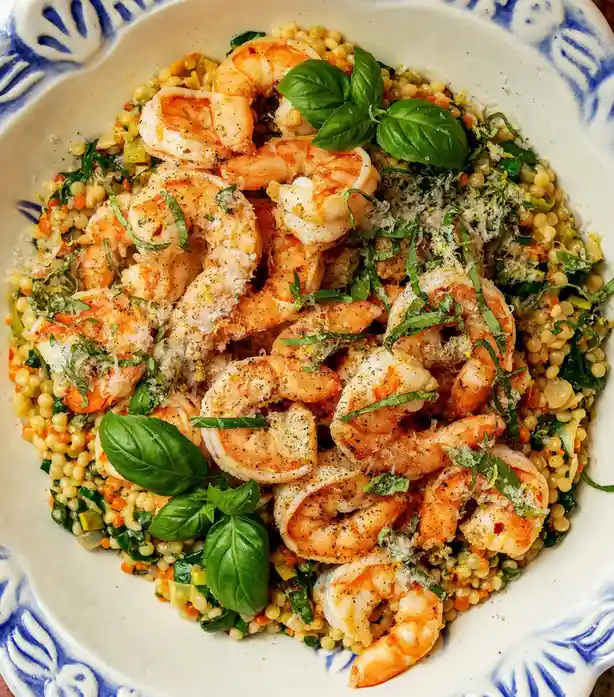 A close-up photo of a white bowl with blue decorative patterns, filled with a colorful dish of cooked shrimp, small pasta or…