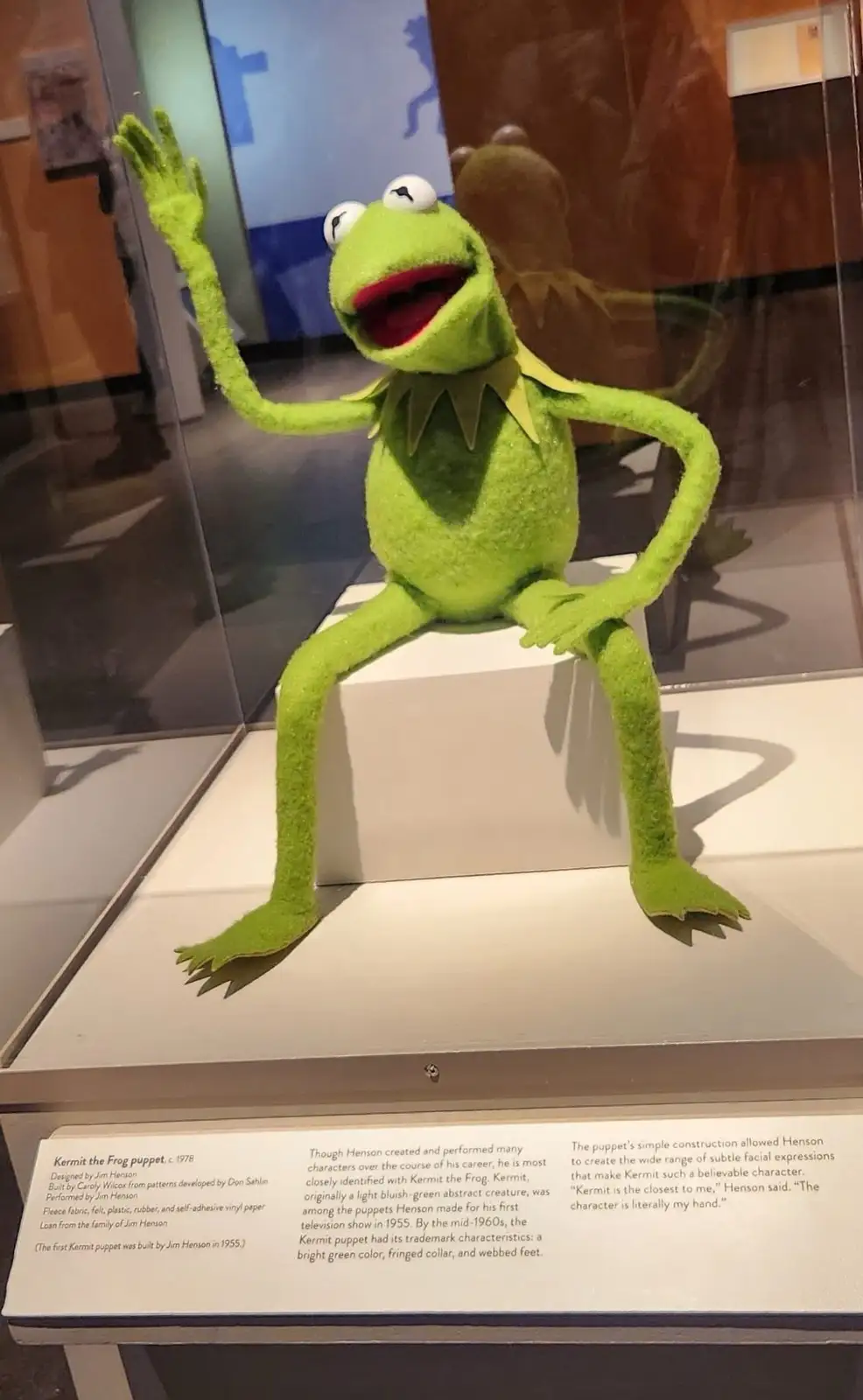 A green felt Kermit the Frog puppet, designed by Jim Henson, is displayed in a museum glass case, sitting on a white…