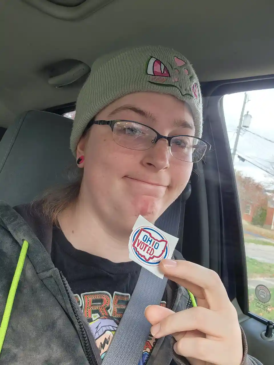 A person wearing glasses and a beanie sits in a car, holding up an 'OHIO VOTED' sticker while smiling. The sticker features…