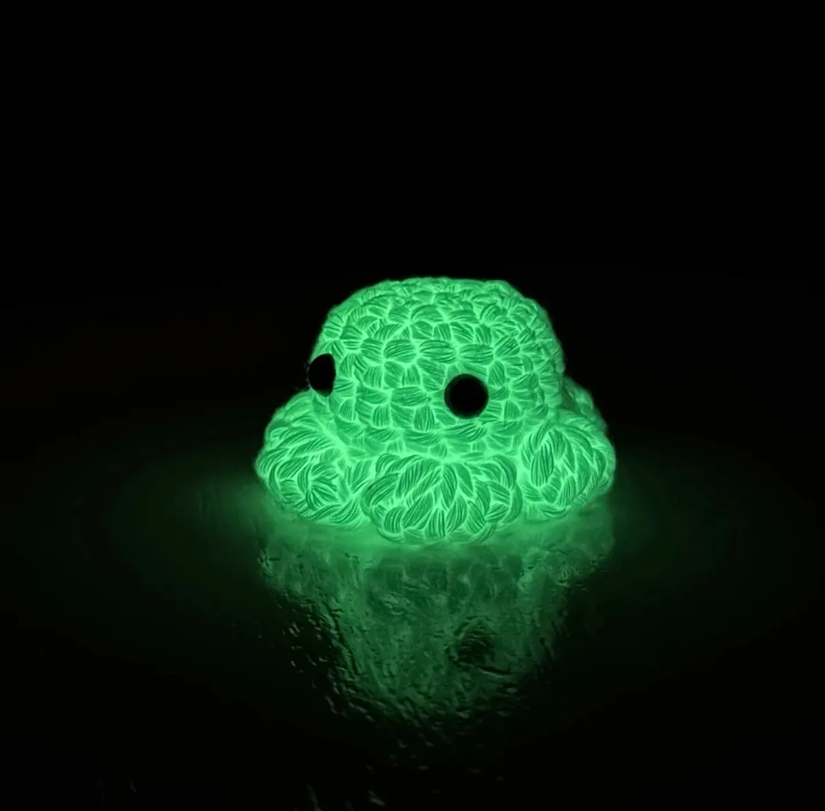 A small, glowing green knitted octopus toy with large black eyes sits on a reflective surface in a dark room, casting a…