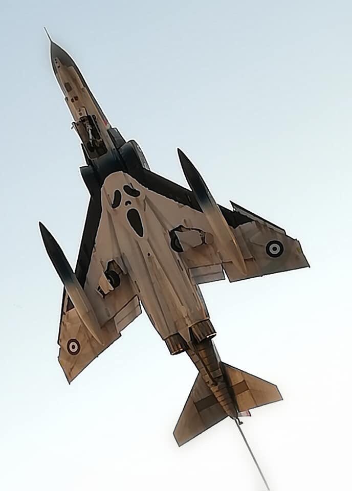 A military jet aircraft is captured from below against a pale sky, its fuselage painted with a large, cartoonish ghost face…