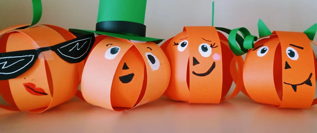 Four handmade orange paper pumpkins with faces are lined up. The first wears sunglasses and red lips, the second wears a…