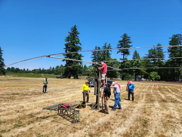 A group of men in a dry grassy field under a clear blue sky are assembling a large, multi-element radio antenna. One man in…