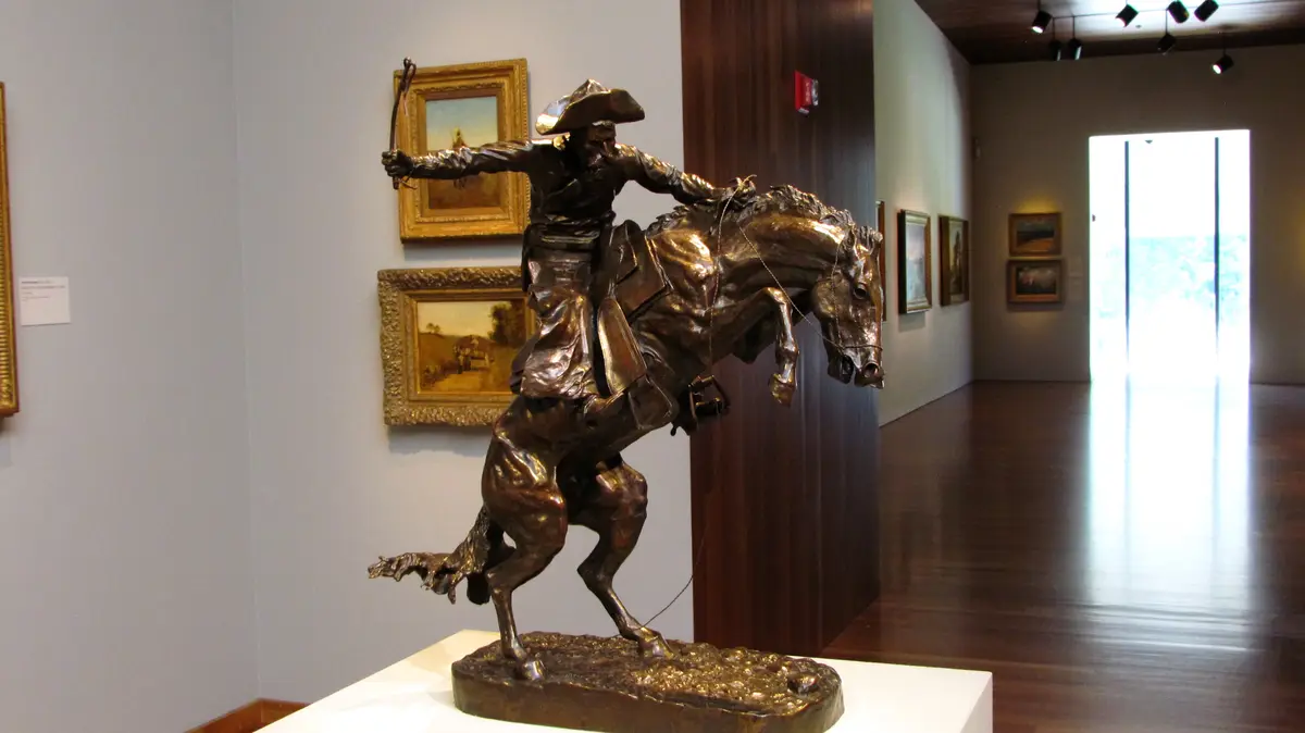 A bronze statue of a cowboy riding a rearing horse is displayed in a museum gallery, surrounded by framed paintings on the…