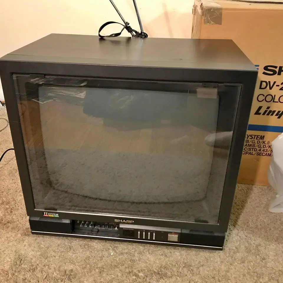 A black Sharp CRT television with a blank, static-filled screen sits on a carpeted floor, with a cardboard box labeled…