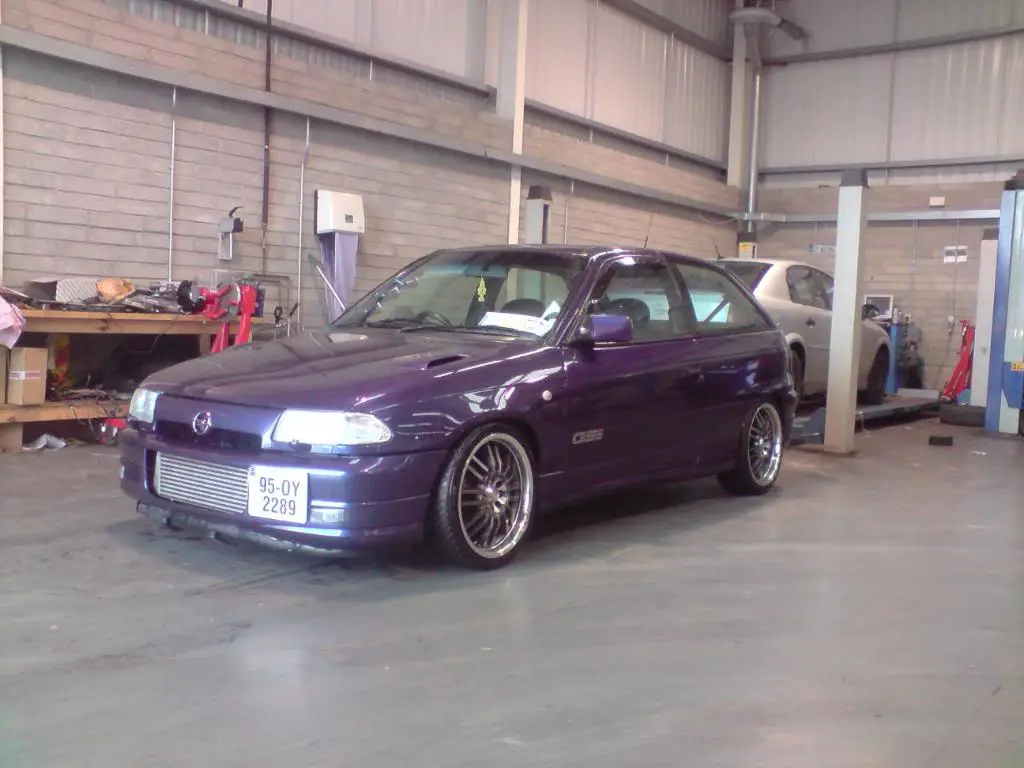 A purple modified Opel Astra GTE is parked inside a garage, featuring aftermarket wheels and a custom front grille. The…