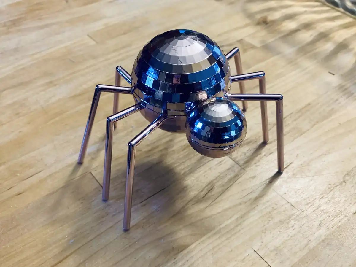 A shiny, metallic spider sculpture with a large blue disco ball for a body and a smaller disco ball for a head, standing on…