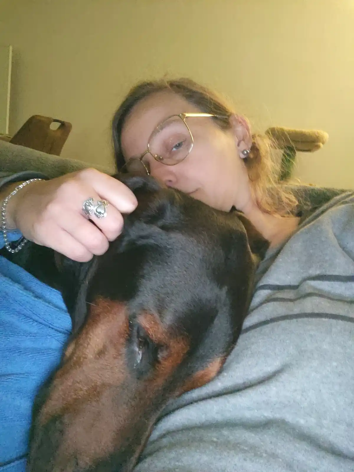 A person with glasses and a grey hoodie is lying down, cuddling a black and tan dog that is resting its head on their chest…
