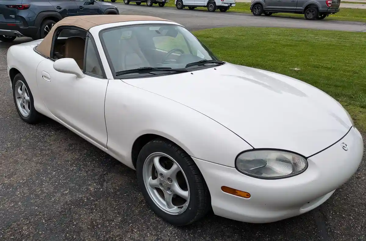 A white Mazda Miata convertible with a tan soft top is parked on asphalt, its roof up, surrounded by other vehicles. The…