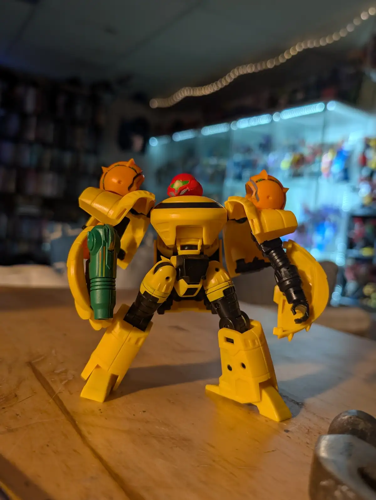 A yellow toy robot with a red head and orange cat-like heads on its shoulders stands on a wooden table, holding a green…