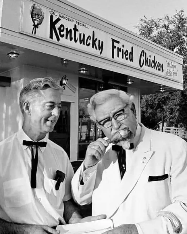 A black and white photo shows Colonel Harland Sanders, founder of Kentucky Fried Chicken, eating a piece of fried chicken…