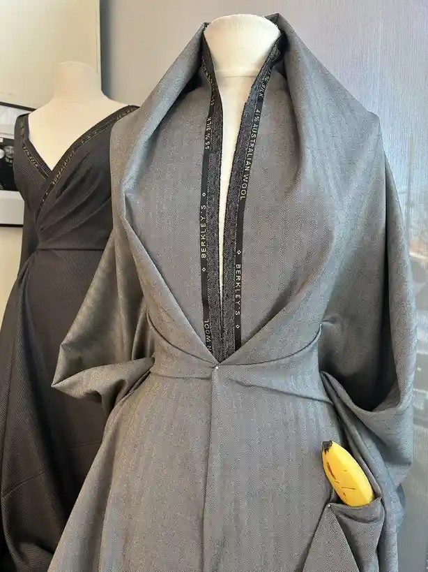 A grey herringbone fabric garment is draped over a mannequin, with a ripe yellow banana visibly sticking out of its pocket…