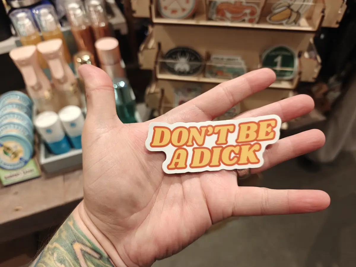 A person's open palm displays a sticker with the text 'DON'T BE A DICK' in bold, orange-yellow letters with a white outline…