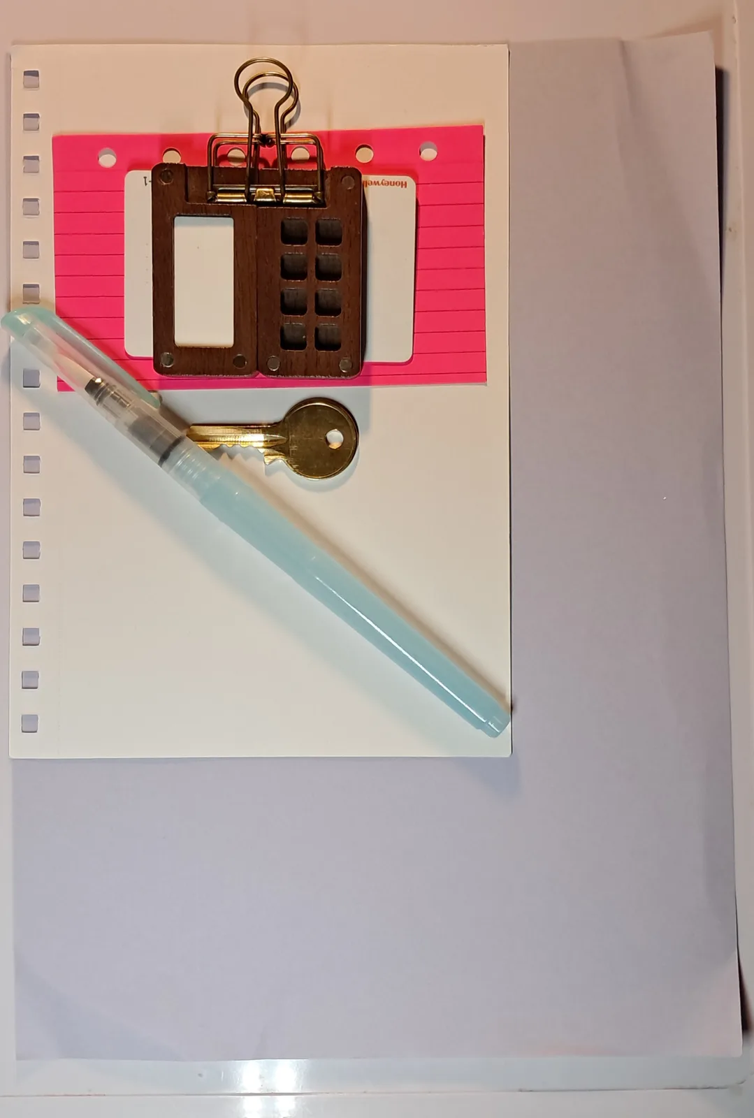 A top-down photo shows a wooden clipboard holding pink lined paper, with a light blue pen and a brass key resting on a sheet…