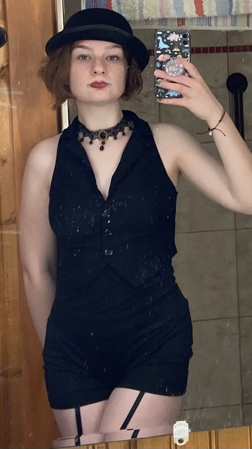 A person with short brown hair wearing a black bowler hat, a black sleeveless romper with buttons, and a black choker…