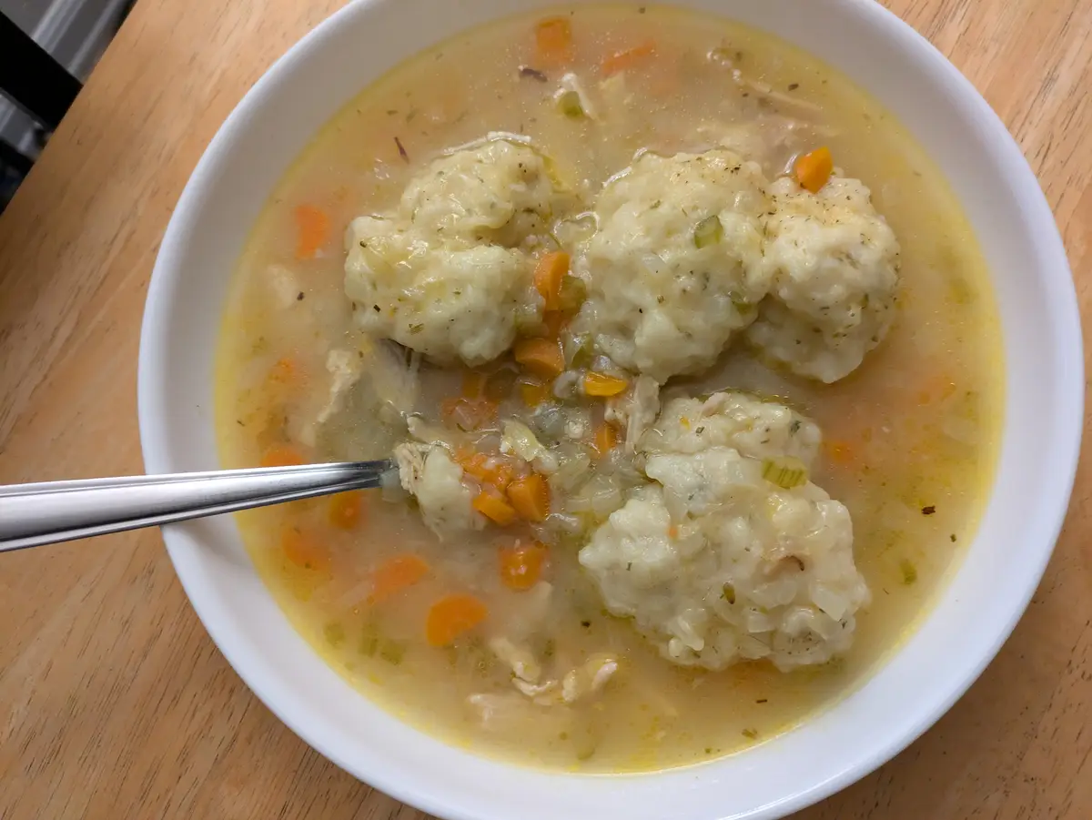 A white bowl filled with chicken and dumplings soup sits on a wooden table, featuring visible dumplings, shredded chicken…