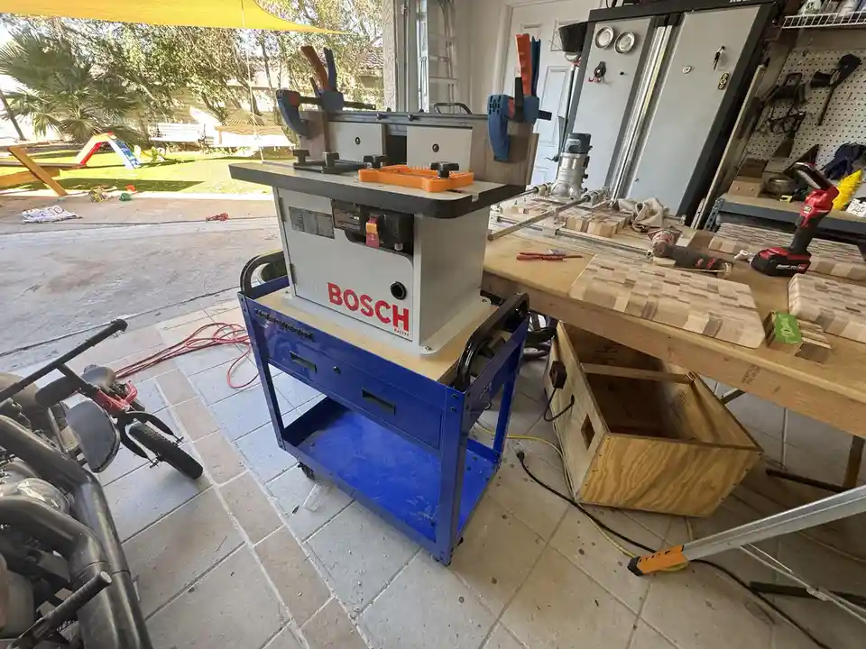 A Bosch table saw sits on a blue rolling cart in a cluttered garage workshop, surrounded by woodworking tools, partially…