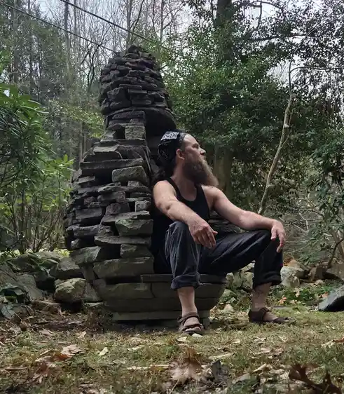 a bearded man with a bandana sits calmly on a large, uniquely stacked stone structure in a wooded area, looking off to the…