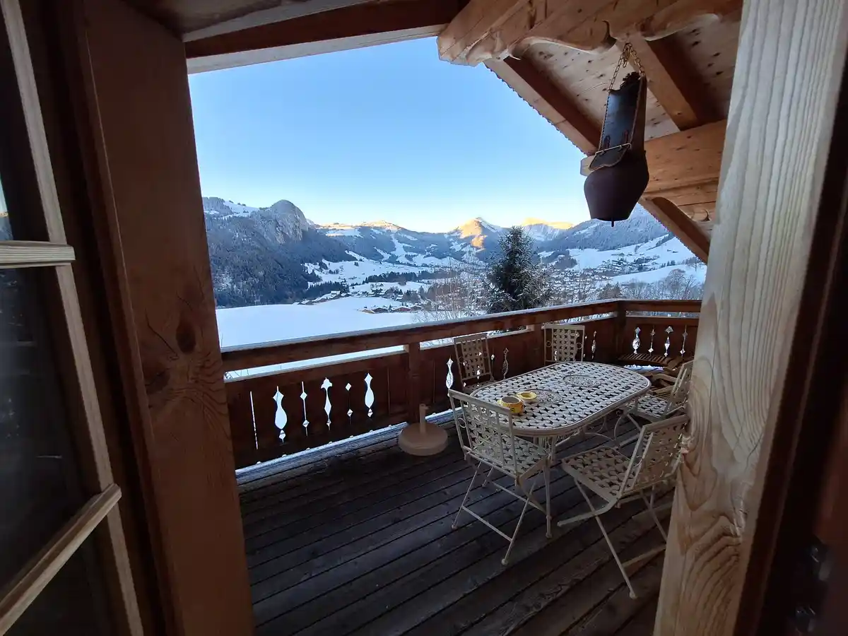 A view from a wooden balcony looking out over a snowy mountain valley with distant peaks under a clear blue sky. The balcony…