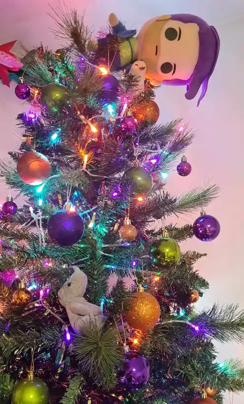 A decorated christmas tree with colorful lights, ornaments, and plush toys, including a large plush character with purple…