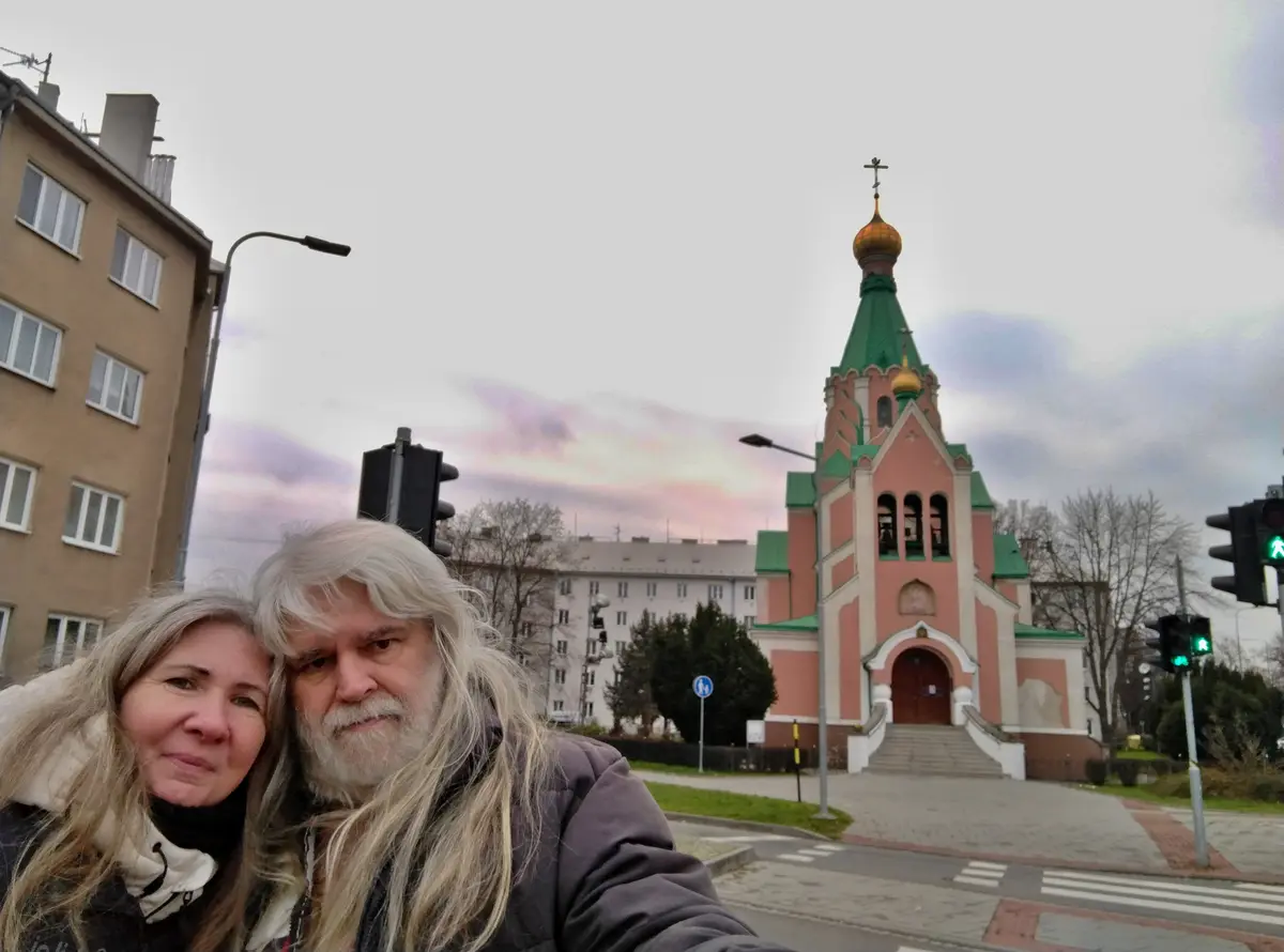 A selfie of an older couple standing on a street corner with a pink and green Orthodox church in the background; the woman…