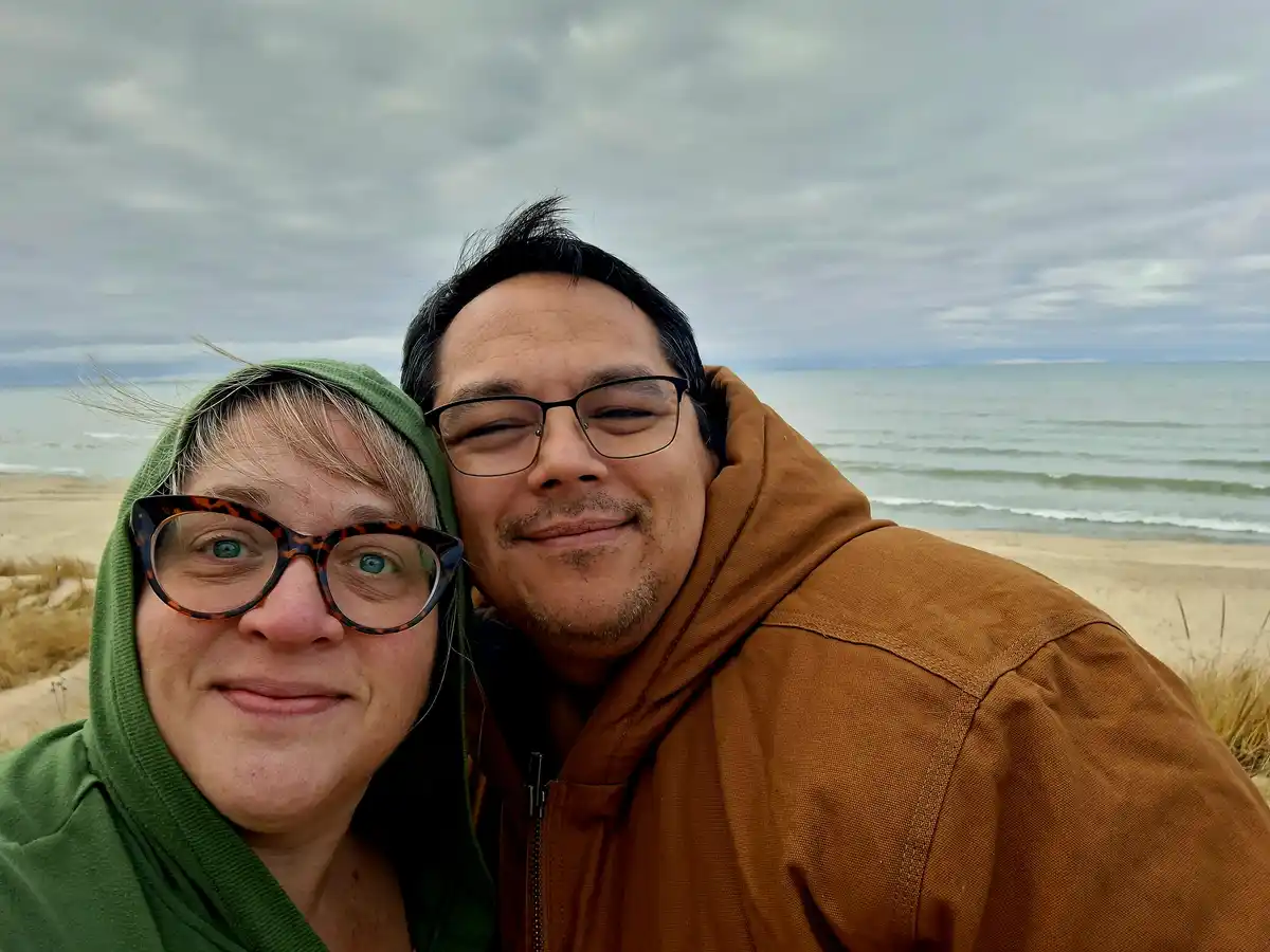 A selfie of a couple on a beach with overcast skies and waves in the background. The woman on the left wears a green hooded…