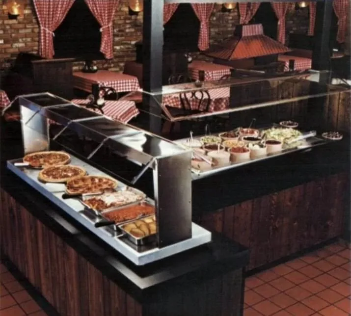 a vintage-style restaurant buffet with stainless steel serving counters displaying pizzas, pasta, and side dishes under…
