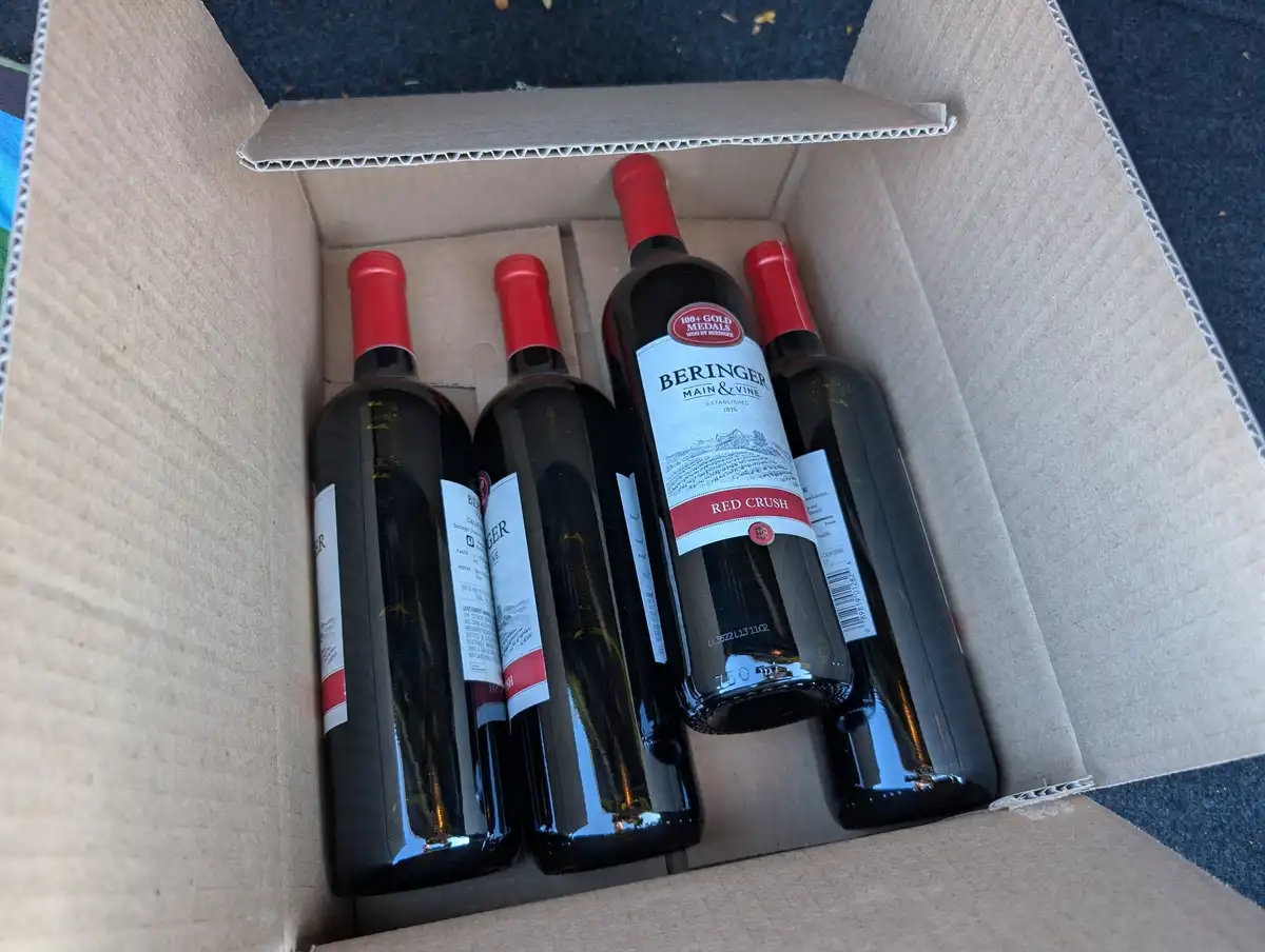 A cardboard box contains four bottles of Beringer Main & Vine Red Crush wine, with one bottle prominently displayed upright…