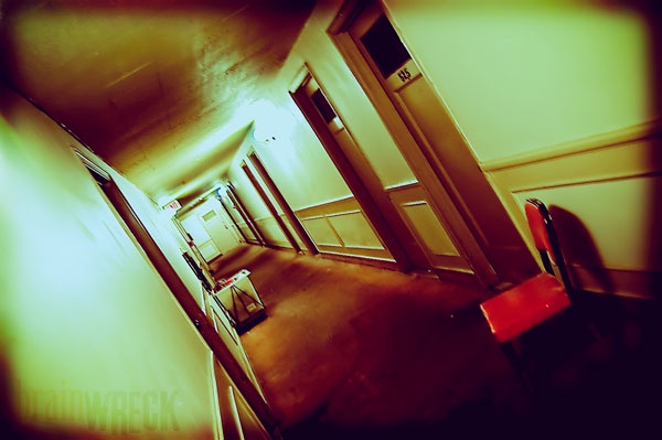 A fisheye lens photo of a dim, yellow-tinted hotel hallway with doors on both sides, one labeled '45', and a red chair in…