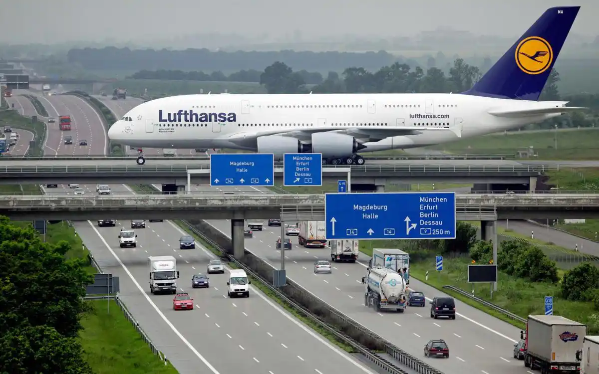 A large Lufthansa airplane is digitally edited to appear as if it's flying directly over a multi-lane highway with cars and…