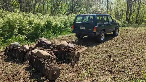 A dark green Jeep Cherokee is parked on a dirt field, attached by a chain to a rusty plow that has large rocks strapped to…