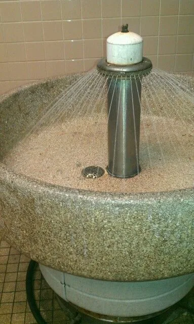 a large, round, speckled stone basin filled with water, featuring a central metal showerhead spraying water outward. The…