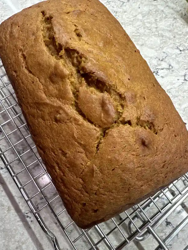 A freshly baked, golden-brown loaf of bread sits on a metal cooling rack atop a speckled countertop. The bread has a…