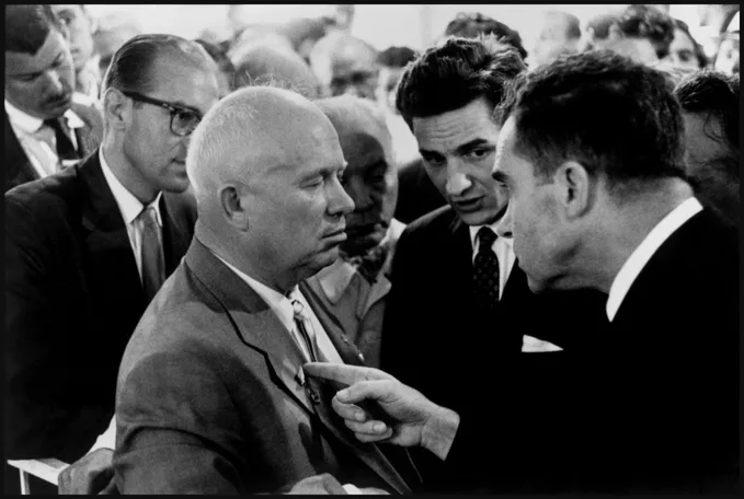 a black and white photo of a man in a suit looking annoyed as another man points at him, surrounded by other people in…