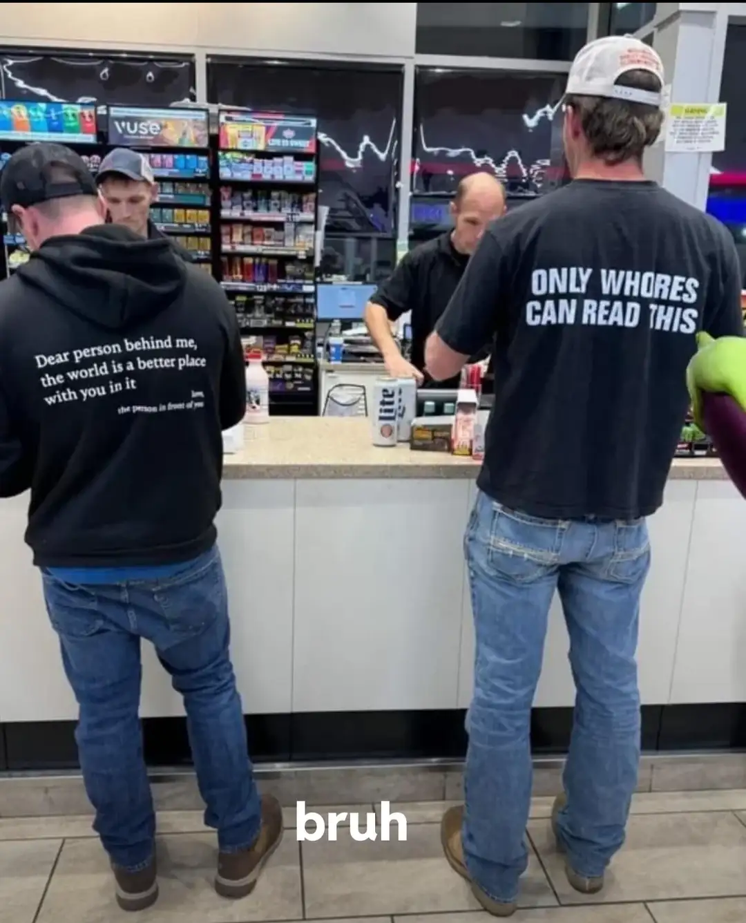 Two men stand at a store counter, viewed from behind. The man on the left wears a black hoodie with the text 'Dear person…