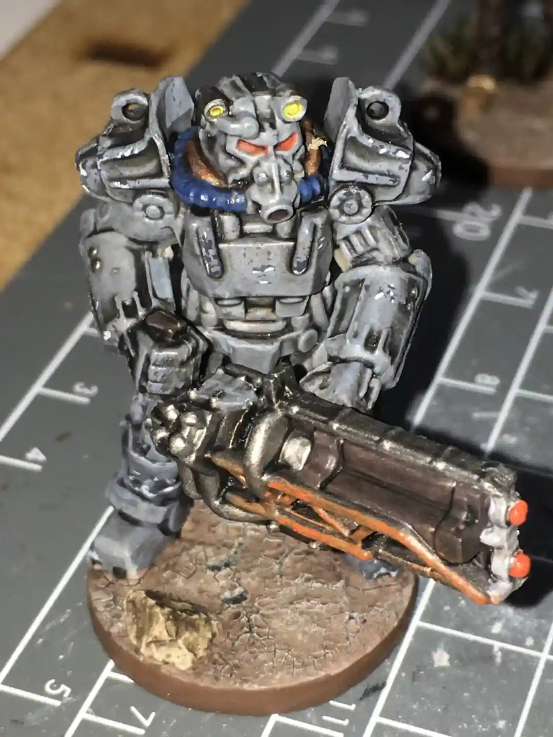 A heavily weathered, grey-painted miniature robot with glowing yellow eyes and red visor, holding a large weapon, stands on…