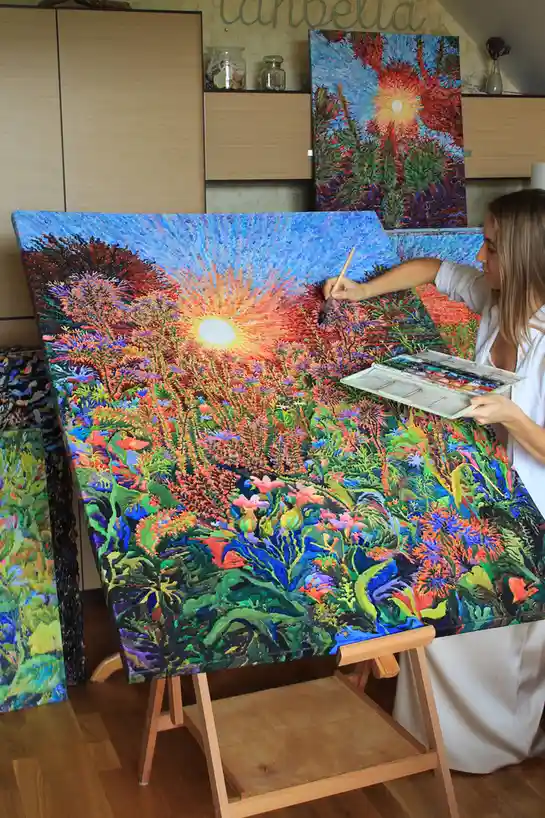 A woman in a white shirt is painting a vibrant, colorful landscape on a large canvas propped on an easel. She holds a…