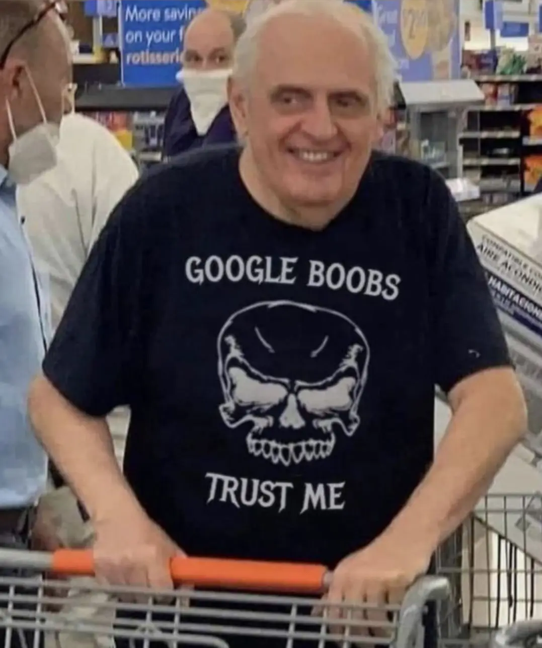 An older man with white hair is smiling broadly while pushing a shopping cart in a store. He is wearing a black t-shirt with…