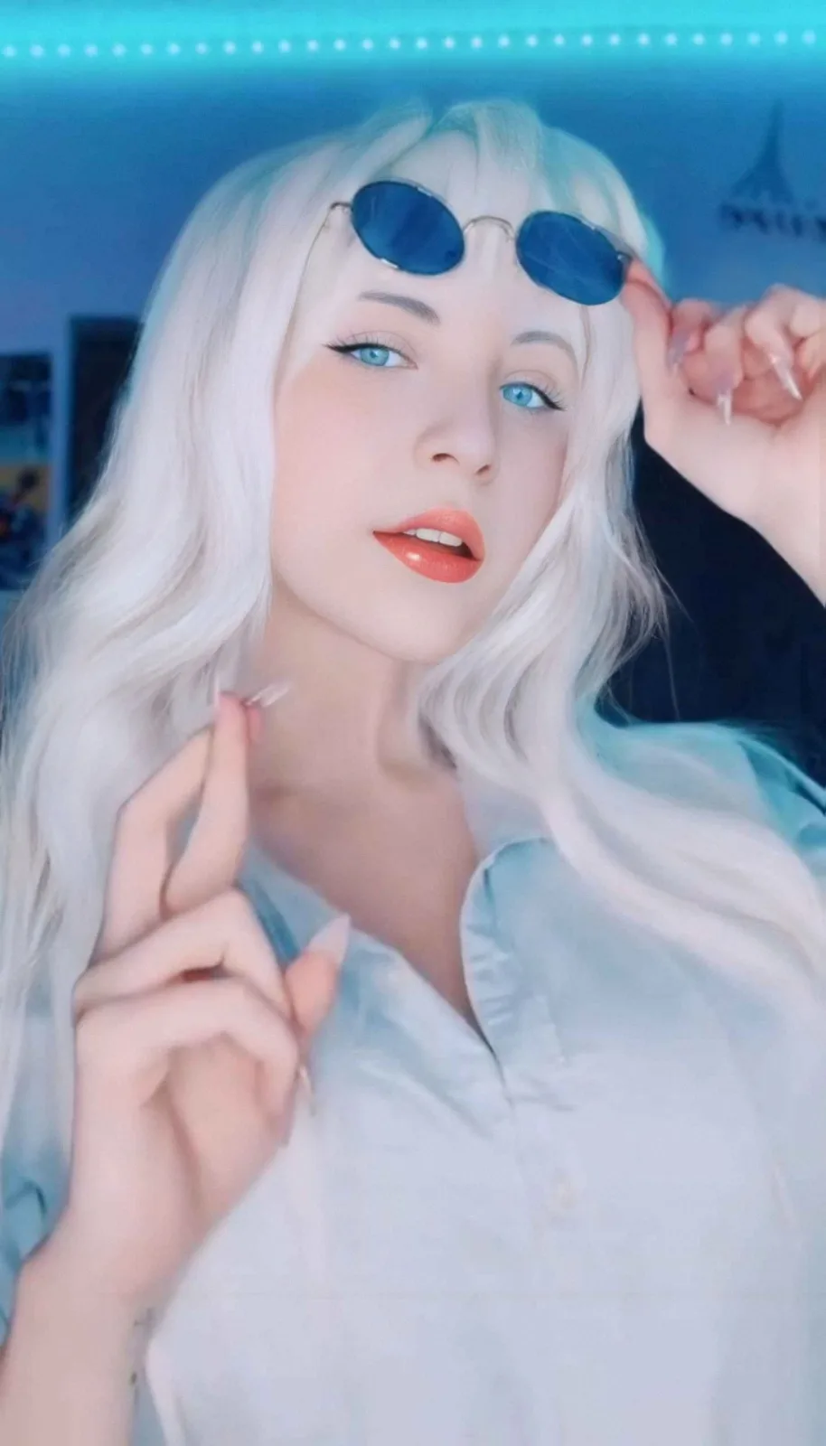 A person with long white hair and blue eyes wears a light blue button-up shirt and has small round sunglasses perched on…