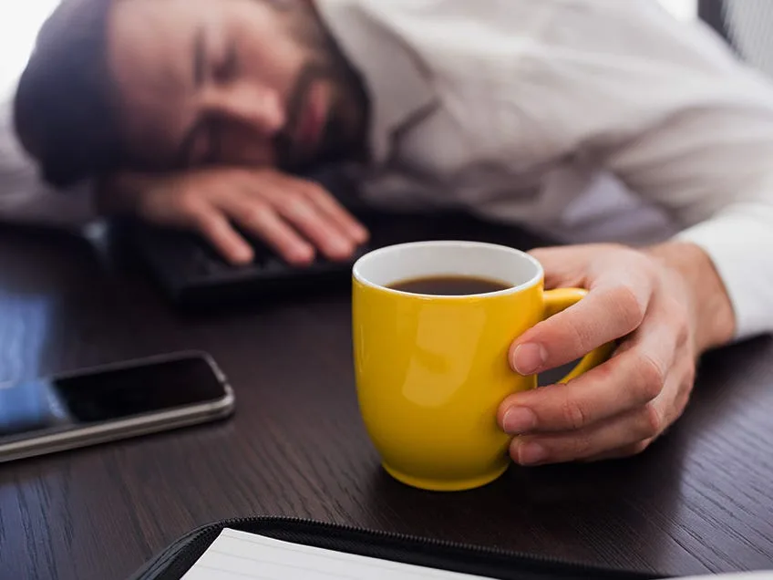 A man with a beard is asleep with his head on a keyboard at a desk, his hand still gripping a bright yellow coffee mug. A…