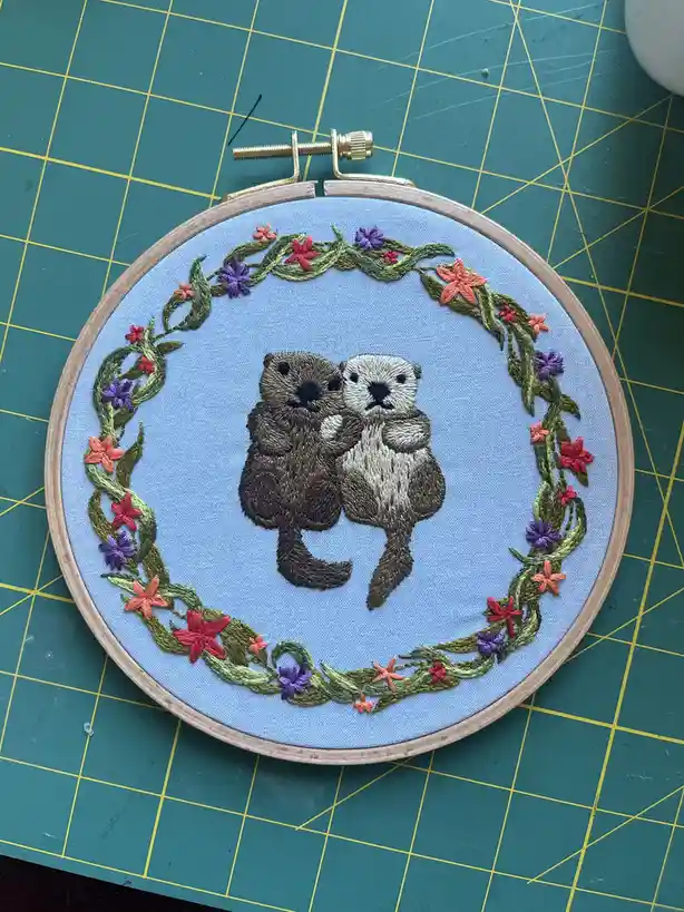 An embroidery hoop holds a light blue fabric with two embroidered otters hugging, surrounded by a colorful floral wreath of…