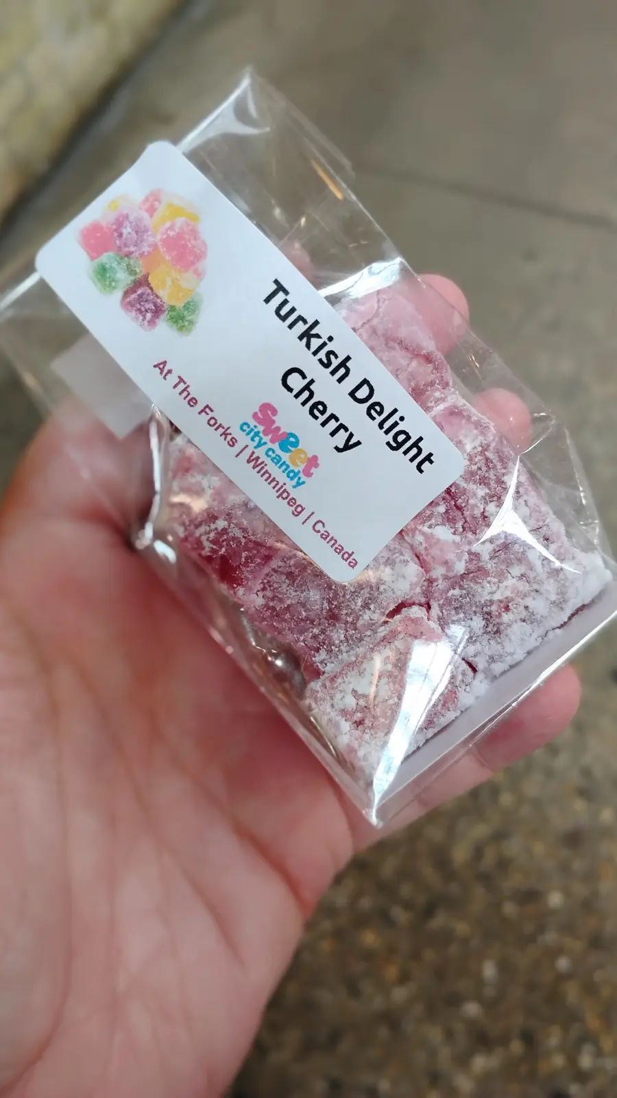 A hand holds a clear plastic bag filled with pink, sugar-coated Turkish Delight candies. The white label on the bag reads…