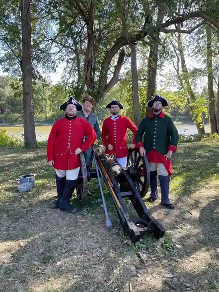 Four men dressed in colonial-era military uniforms stand around a cannon in a wooded area near water. Three wear red coats…