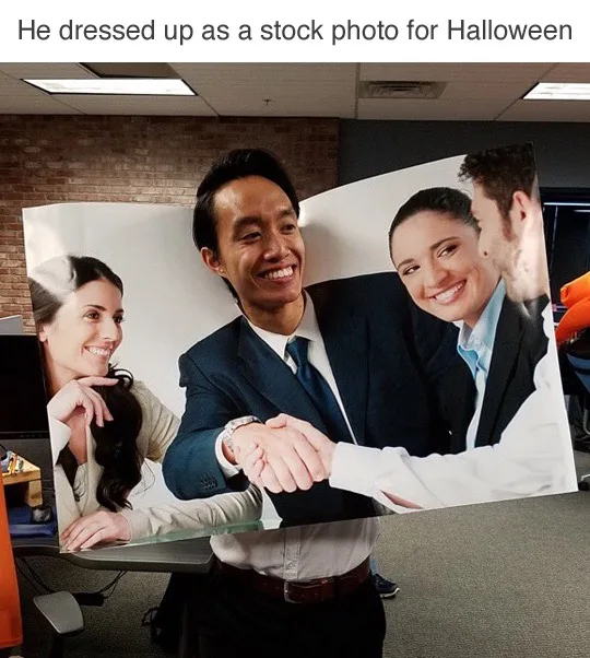 A man in an office is wearing a large printed stock photo as a costume, depicting two people shaking hands while a third…