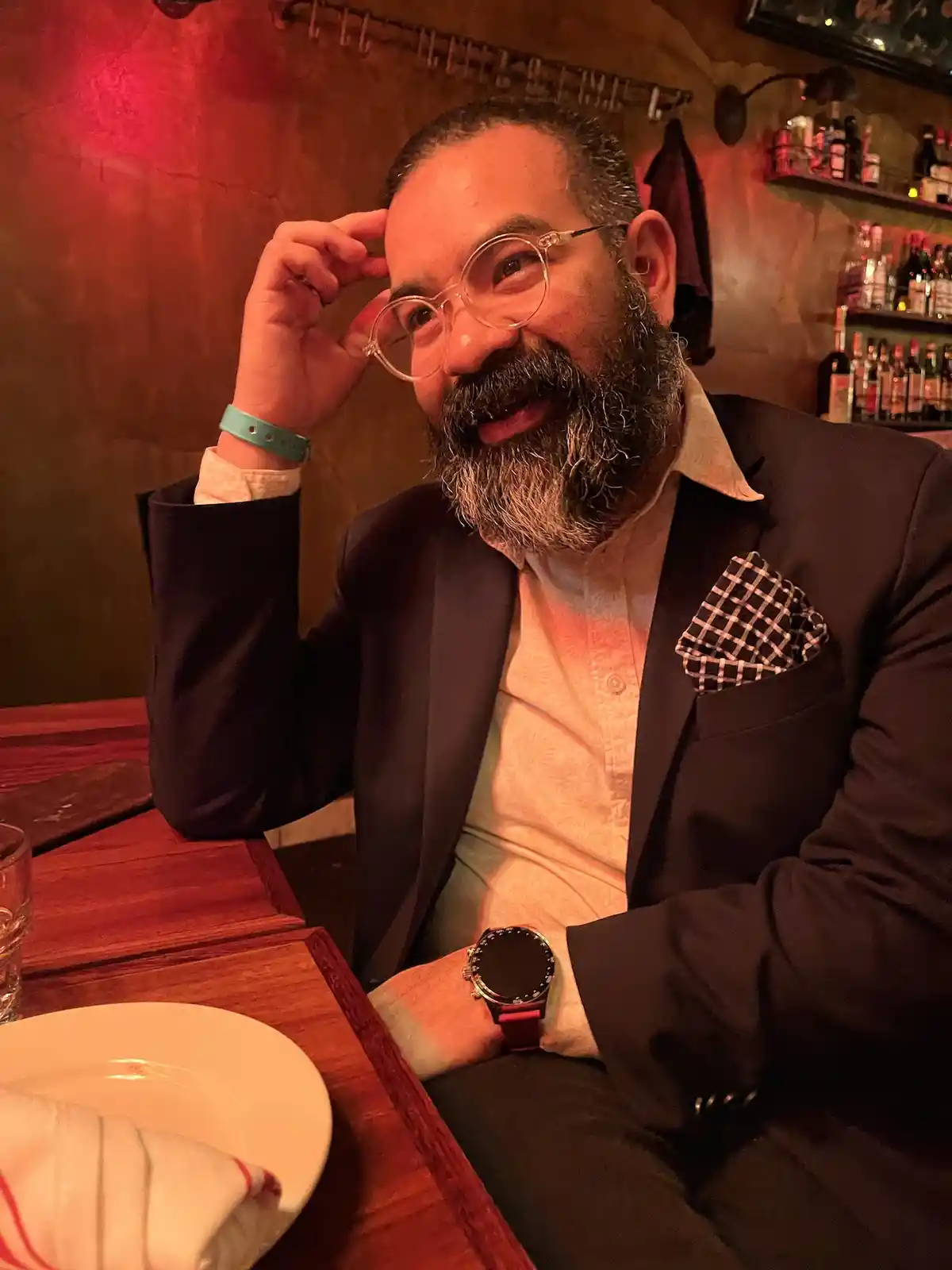 A man with a beard and round glasses sits at a restaurant table, smiling and adjusting his glasses with one hand. He wears a…