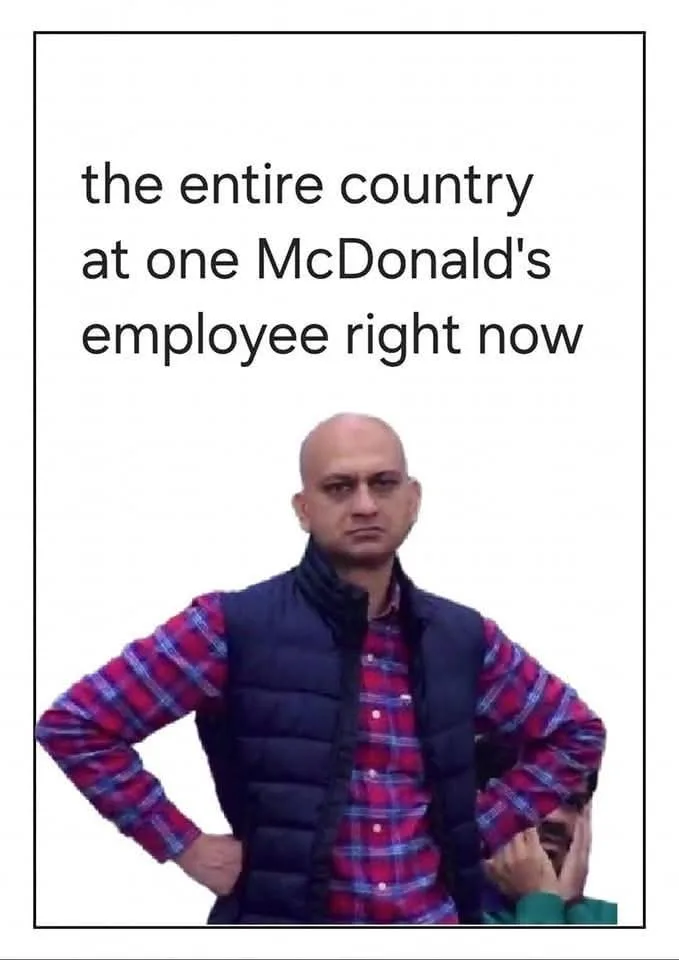 A bald man in a red and blue plaid shirt under a dark vest stands with hands on hips, looking sternly at the camera. Behind…